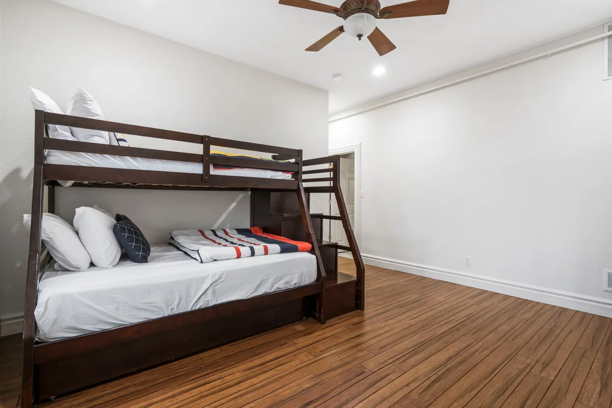 A cozy kids' bedroom with a sturdy wooden bunk bed, ceiling fan, and warm wooden flooring. The space is clearly designed for children or young guests, encouraging family togetherness and playful sleepovers.