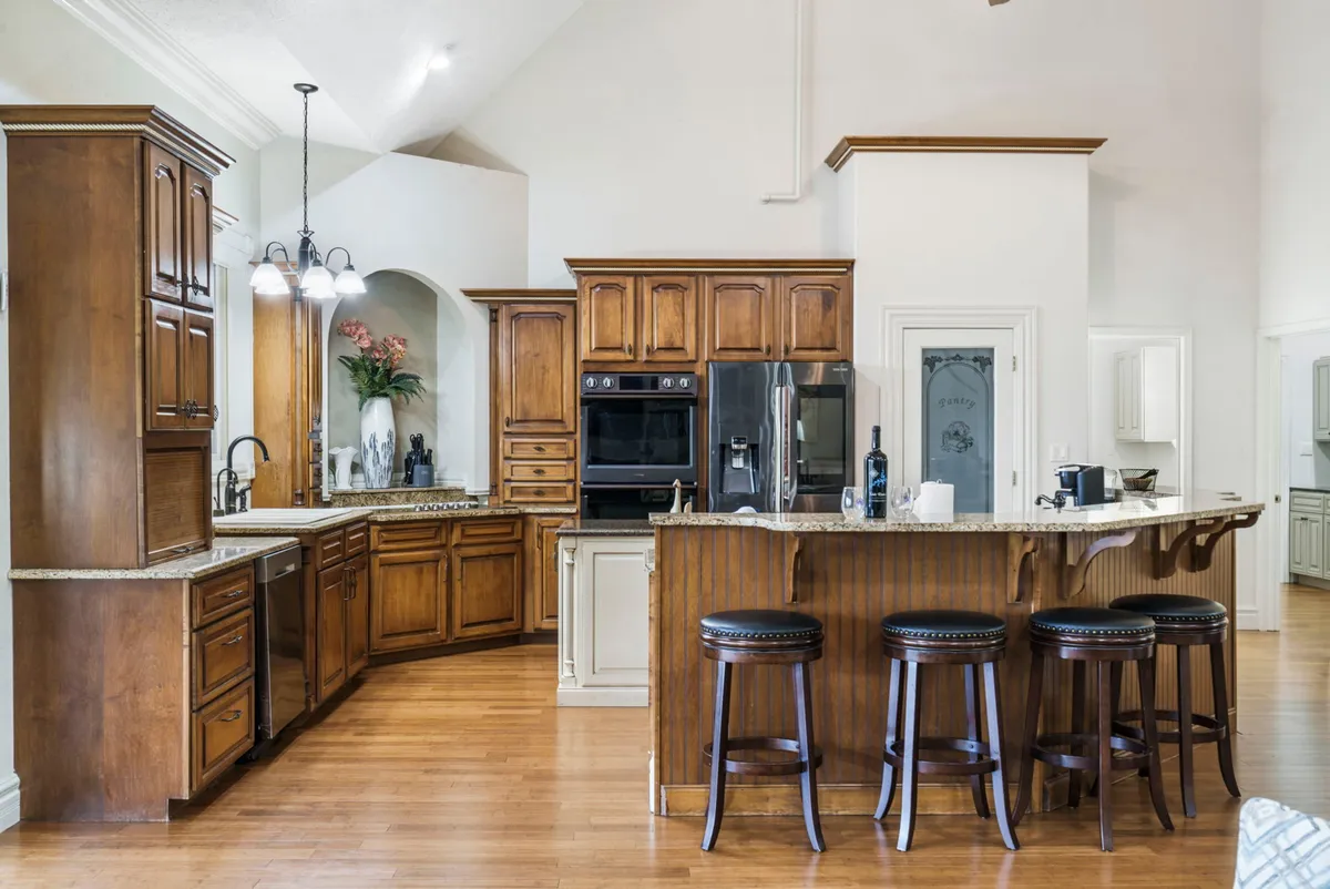 A close-up of the kitchen island with bar stools, wood cabinetry, granite countertops, and modern appliances. The space exudes warmth and is designed for cooking, casual dining, and entertaining.