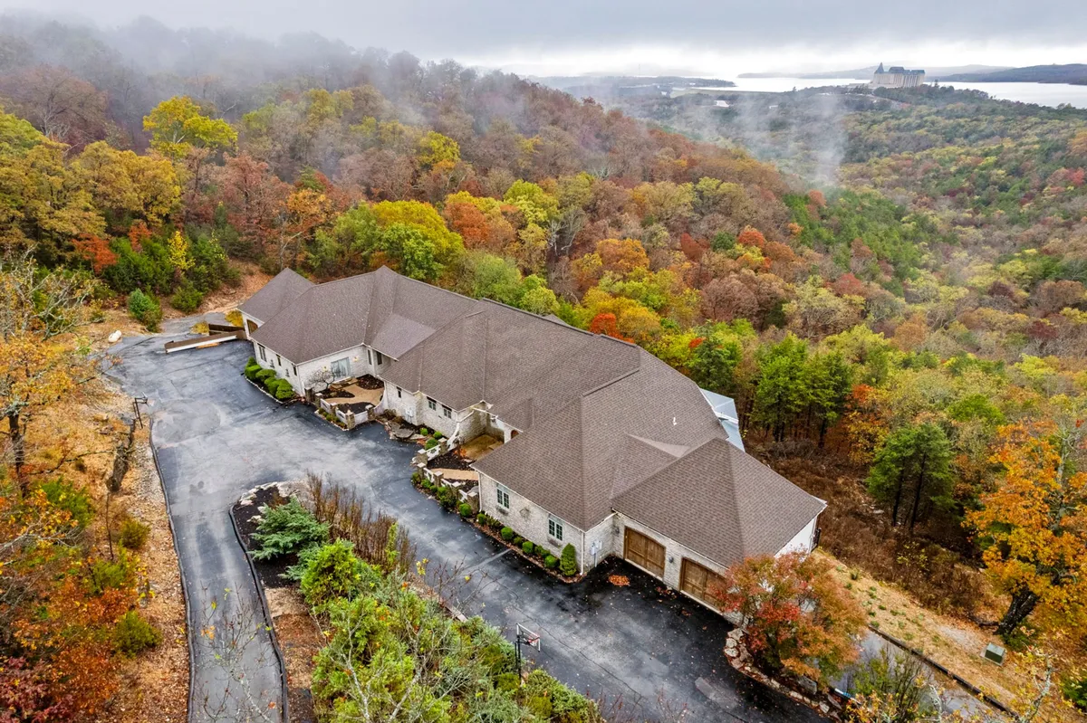 The image showcases the exterior of a spacious townhouse nestled among mature trees, with a welcoming driveway curving toward the front of the home. The tranquil natural setting is highlighted by colorful foliage, suggesting a peaceful retreat, and hints at outdoor spaces perfect for gatherings or relaxation.